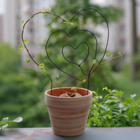 A potted plant with a heart-shaped wire trellis made of black wire, featuring green leaves and a small plant growing on it, placed on a windowsill with a blurred background of greenery.